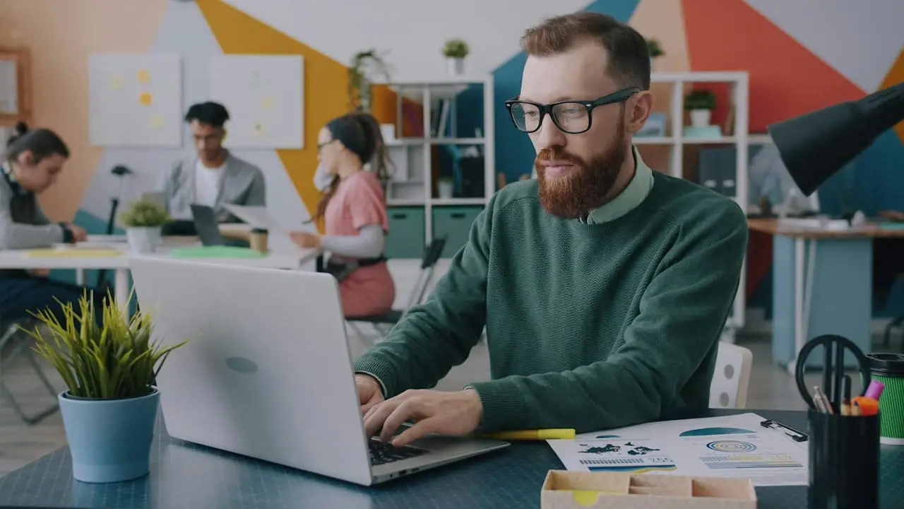 Un professionnel au travail sur son ordinateur, ses documents chiffrés posés à côté de lui